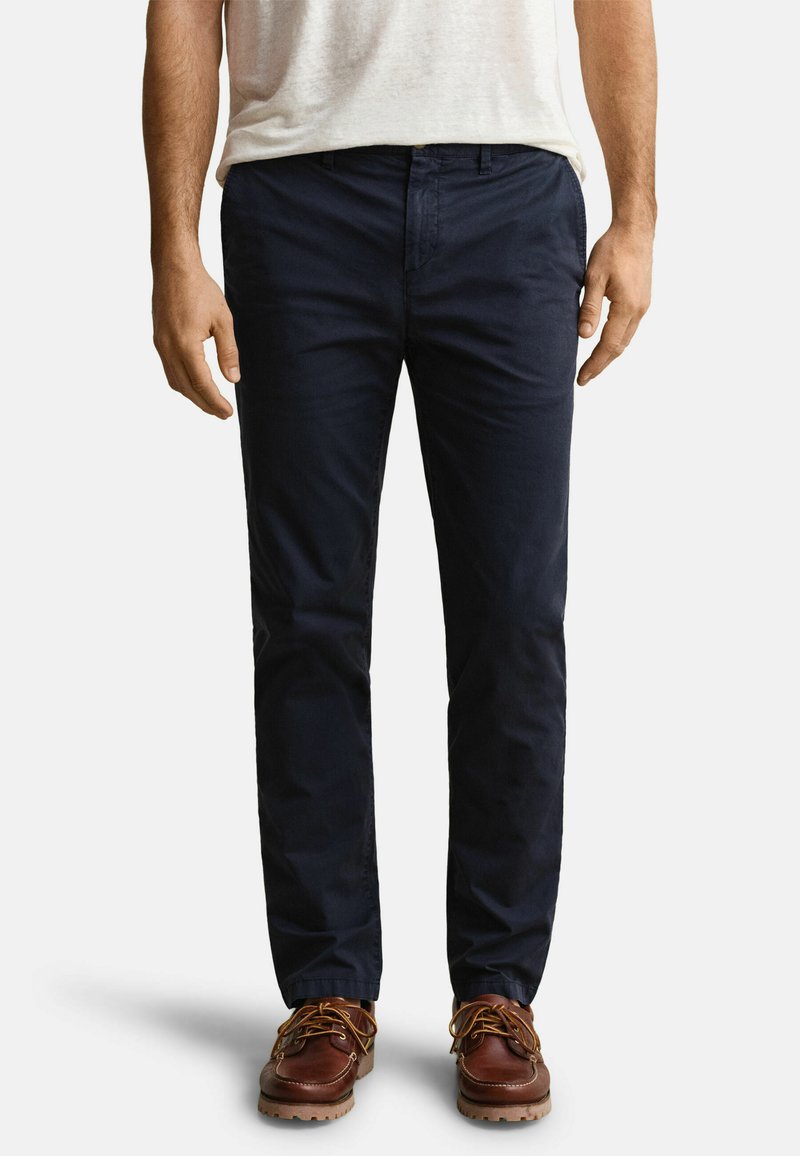 Hombre con pantalones chinos azul marino oscuro, zapatos marrones de piel con cordones y camisa de manga corta beige claro, de pie frente a un fondo liso.