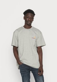 Gray cotton t-shirt with short sleeves, round neckline, and a small orange and red Carhartt logo on the chest, paired with dark denim jeans.