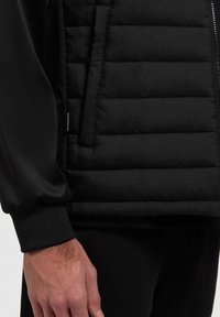 Zwarte gewatteerde vest met een quilted afwerking, voorzien van een gladde stofpaneel, ritsluiting en ribgebreide manchetten voor extra detail.
