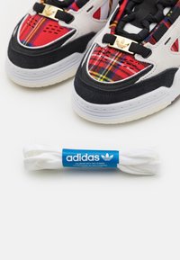 Zapatillas Adidas con parte superior de cuadros rojos, amarillos y negros, detalles en ante negro y suela de goma blanca. Incluye cordones blancos en el embalaje.