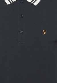 Navy polo shirt with a textured fabric, featuring a white striped collar, three black buttons, and an embroidered yellow logo on the chest.