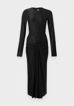 Moteris dėvi juodą midi suknelę ilgomis rankovėmis su raukšlėtu mazgo detaliu juosmenyje, derina juodus aukštakulnius sandalus su čiurnos dirželiais.