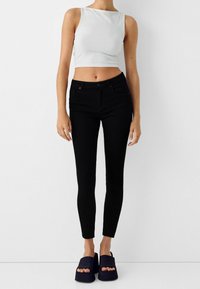 Mujer vestida con una blusa sin mangas blanca, jeans ajustados negros y sandalias de plataforma negras, de pie contra un fondo blanco liso.