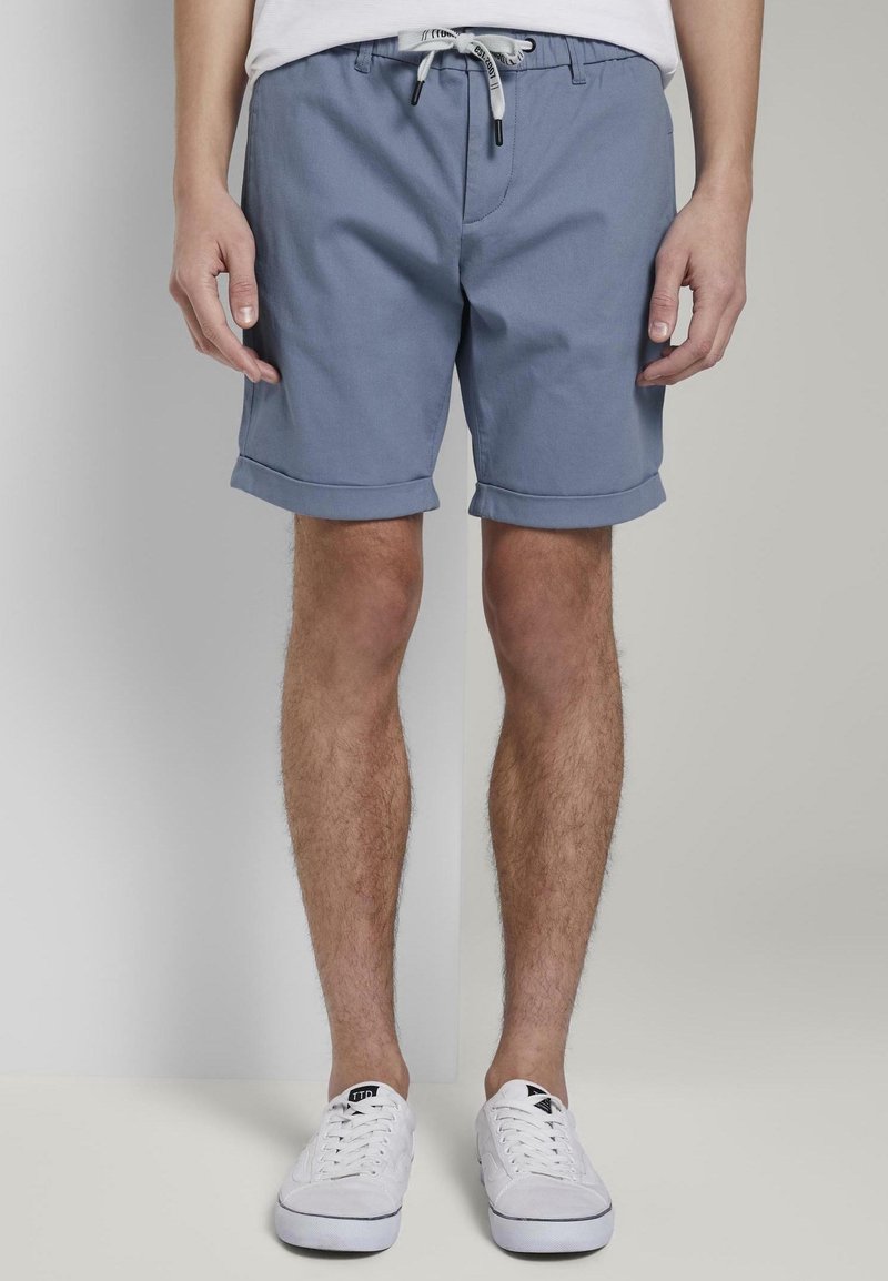 Pantalones cortos de algodón azul claro con cintura ajustable, puños doblados y un bolsillo frontal. Llevados con zapatillas blancas.