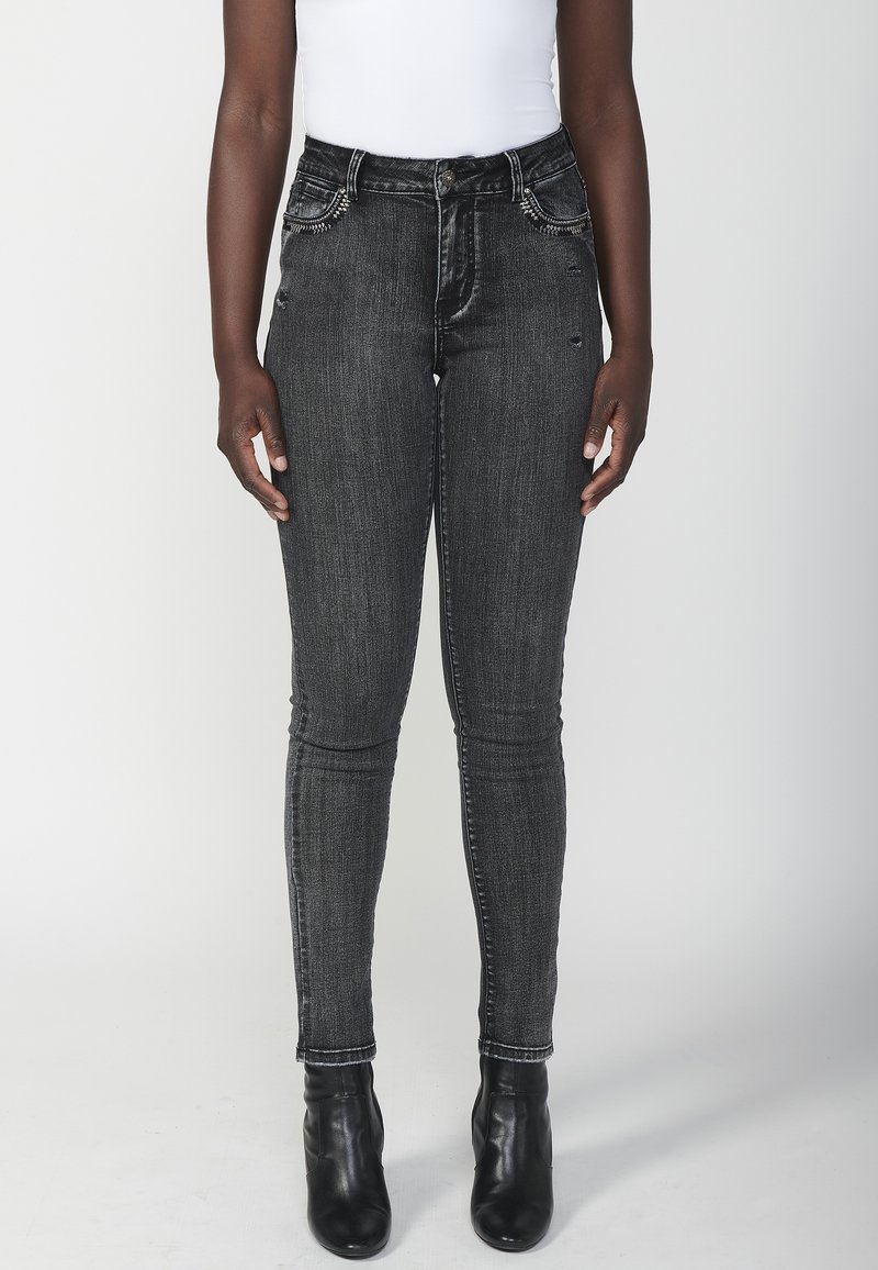 Črne ožje kavbojke s obrabljenim videzom, petimi žepi in dekorativnim šivanjem spredaj. Nosi se jih z črnimi gležnjarji.