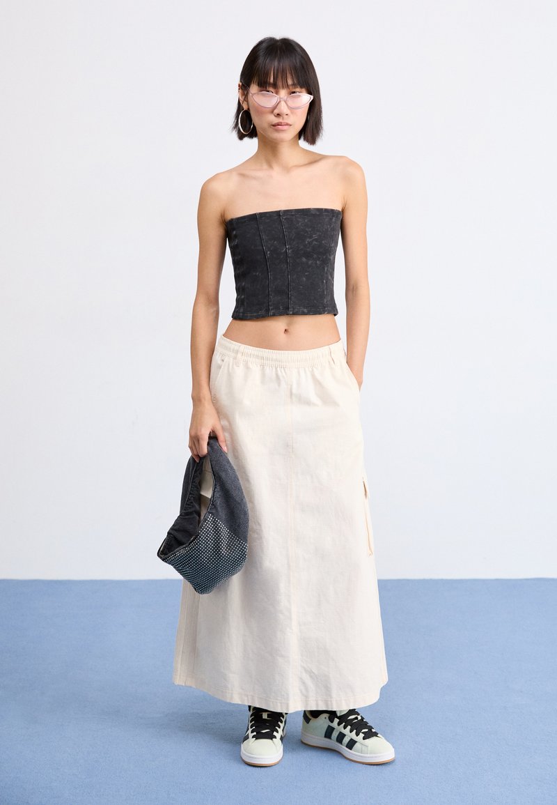 Top senza spalline in denim nero, gonna lunga crema con tasche laterali, borsa grigia a motivo e sneakers alte bianche e nere.