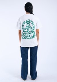 Bílý tričko s velkým zeleným květinovým designem mírového znamení na zádech, obklopeným textem. Vzhled doplněný modrými zvonovými džíny.