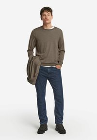 Brauner Langarmpullover mit rundem Halsausschnitt, kombiniert mit dunkelblauen Jeans. Die Person hält einen passenden Pullover und trägt dunkle Schuhe.