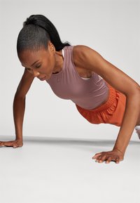 Completo sportivo composto da un top senza maniche di colore lavanda aderente e pantaloni corti arancioni. Il soggetto esegue un push-up su una superficie piana.