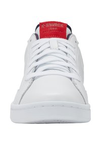 Zapatilla de cuero blanca con cordones planos blancos, acentos rojos y marinos, y suela de goma texturizada. Presenta el logo en la lengüeta y en el lateral.