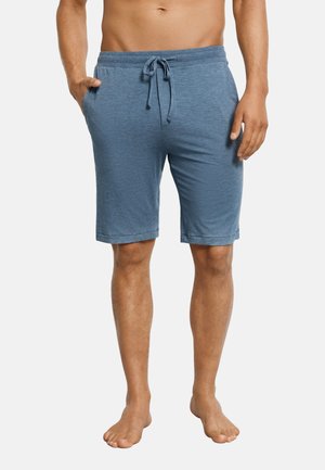 Mężczyzna ubrany w niebieskie spodenki z troczkiem i kieszeniami, stojący boso na białym tle.