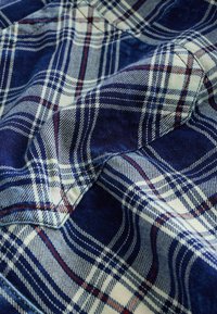 Blauw-witte geruite stof met een gestructureerde weefsel, met rode en donkerblauwe accenten, die opgevouwen secties toont die diepte creëren.