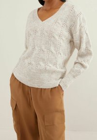 Pull en maille torsadée gris clair avec un col en V et un ourlet côtelé. Présente des motifs texturés et des manches longues. Porté avec un pantalon cargo beige.