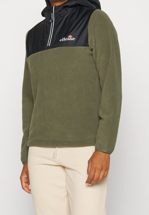 Mand iført olivengrøn fleece-pullover med sort hætte og hvid lynlås, kombineret med off-white bukser.