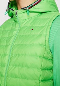 Gilet imbottito verde con chiusura a zip, dotato di cappuccio e accenti in rosso e blu navy a contrasto. Materiale leggero, texture trapuntata.