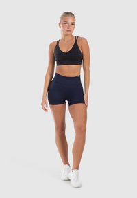 Zwarte sportbh met een V-hals en geplooide details, gecombineerd met hooggetailleerde marineblauwe shorts. Model draagt witte sneakers, tegen een effen achtergrond.