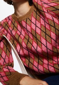 Personne portant un pull à losanges marron et rose, assise avec un bras levé, montrant un tricot texturé et des poignets côtelés.
