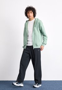 Um hoodie verde claro com fecho, sobre uma T-shirt branca, combinado com calças largas pretas com um logótipo e sapatilhas cinzas com detalhes azuis.