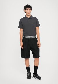 Mørk grå polo skjorte med krage og rund logo på brystet, parret med svarte knelange shorts og svarte treningssko med hvite detaljer.