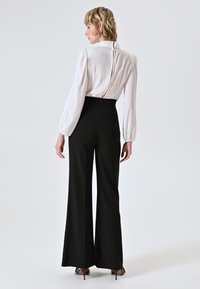 Blusa bianca con maniche a sbuffo, chiusura con bottoni sul retro, abbinata a pantaloni neri a vita alta e svasati. Texture lisce e vestibilità sartoriale.