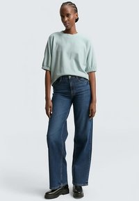 Hellgrünes, kurzärmeliges Oberteil mit gerafften Bündchen, kombiniert mit dunkelblauen, hoch taillierten Jeans. Modell steht mit entspannten Händen, schwarze Stiefel.