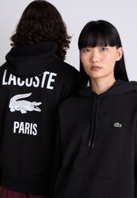 Juodi džemperiai su kapuci turi priekinę kišenę, mažą žalią krokodilo logotipą ir didelį baltą užrašą "LACOSTE PARIS" ant nugaros.