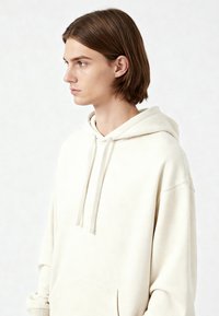 Hoodie color crema realizzato in tessuto morbido, con tasca frontale, cordini e una vestibilità comoda con una texture delicata e dettagli petrosi.