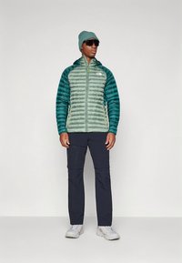 Žalia ir mėlyna šilta striukė su užtrauktuku priekyje, derinama su tamsiomis kelnėmis ir šviesios spalvos batais. Modelis dėvi kepurę ir akinius nuo saulės.