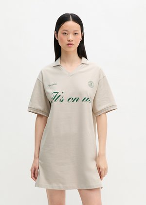Jonge vrouw die een beige polojurk met korte mouwen draagt met groene tekst "It's on us" en logo op de borst, staand tegen een witte achtergrond.