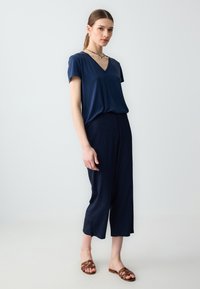 T-shirt bleu marine à col en V avec des manches courtes, associé à un pantalon bleu marine texturé et coupé, et des sandales à lanières marron, sur un fond clair.