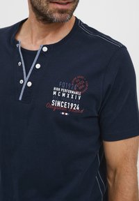 Tamsiai mėlynas medvilnės polo marškinėliai su trumpomis rankovėmis, mygtukų apykakle, baltais kontrastingais siuvinėjimais ir raudonais bei baltais atspaudais.
