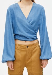 Haut cache-cœur en denim bleu clair à manches longues bouffantes et décolleté en V profond, associé à un pantalon taille haute couleur sable.