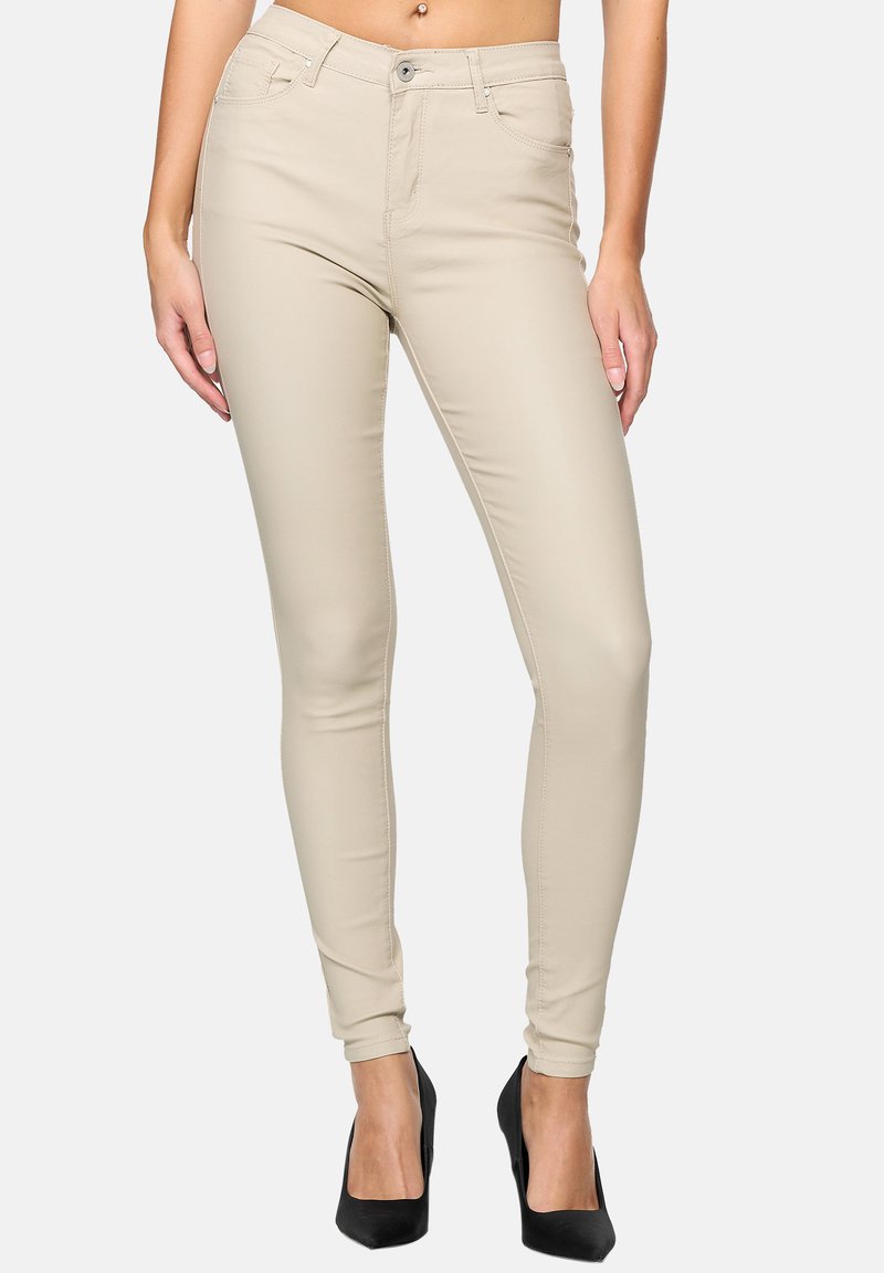 Jean skinny beige avec une texture lisse, design à cinq poches et fermeture à bouton sur le devant. Porté avec des talons hauts noirs.