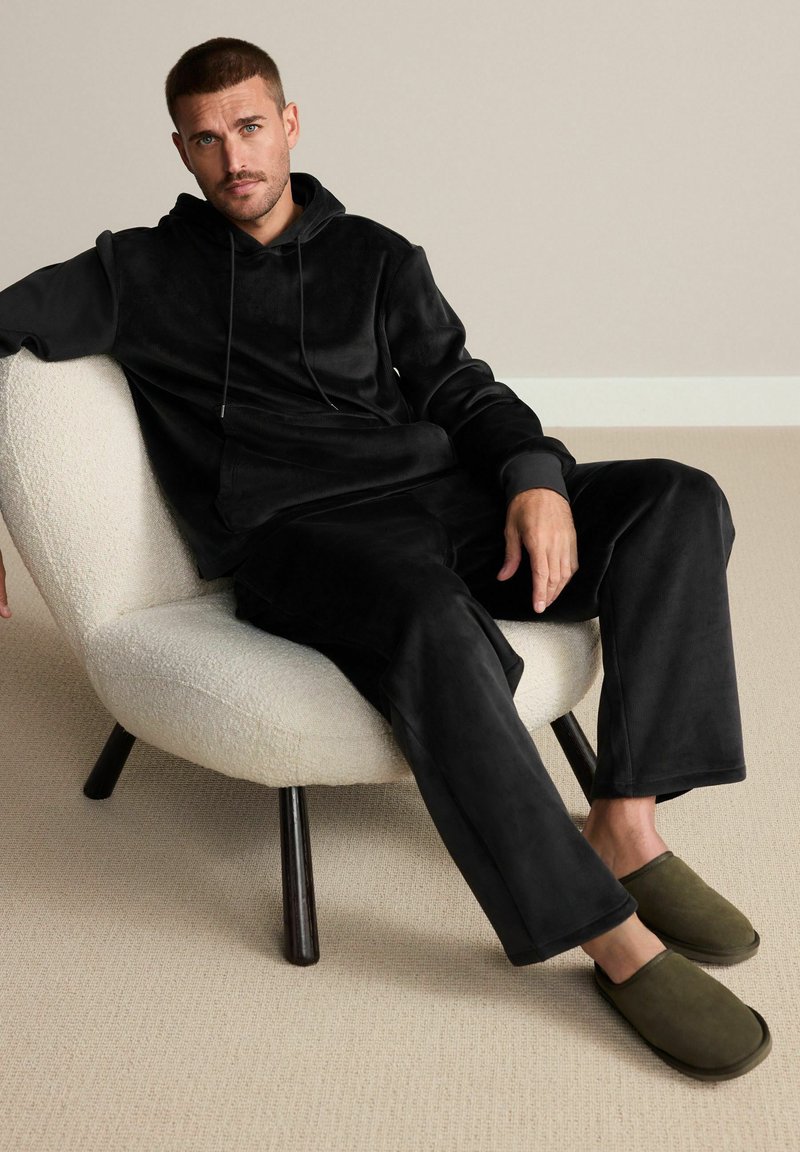 Black fleece hoodie and pants set, soft texture, relaxed fit. Olive green slip-on slippers. Light-colored, round chair with black legs.