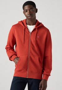 Felpa rossa con zip, realizzata in tessuto morbido, caratterizzata da un cappuccio con coulisse, tasche a marsupio, polsini a costine e una piccola etichetta con logo sul petto.