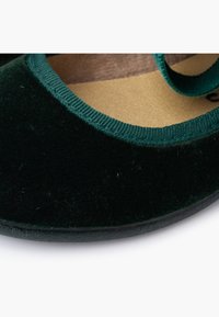 Zapato plano de terciopelo verde con punta redondeada, que presenta una superficie texturizada y un ribete verde contrastante a lo largo de la abertura. Suela de goma negra.