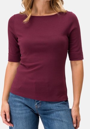 Woman wearing a fitted burgundy elbow-sleeve top and blue jeans, standing against a plain light background.