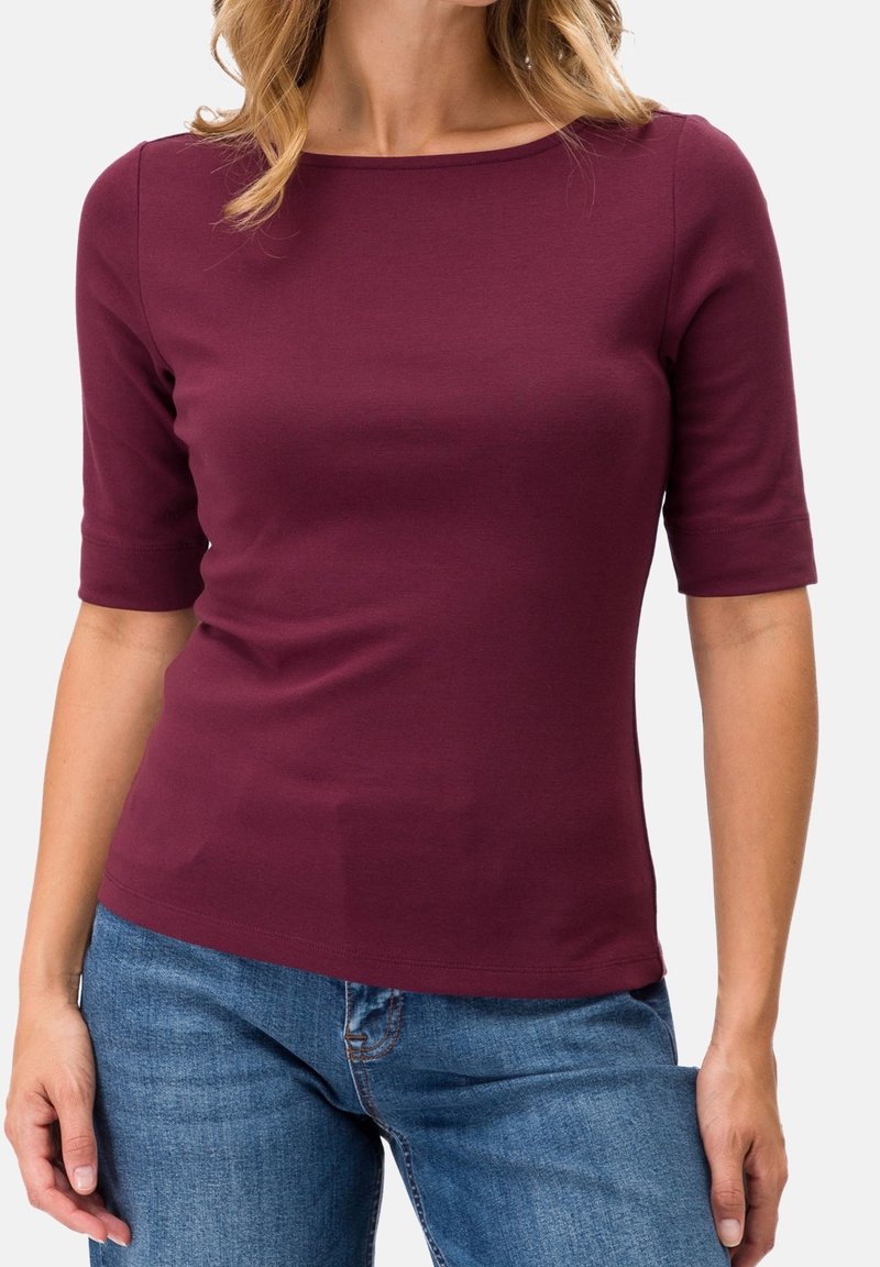Femme portant un haut bordeaux ajusté à manches aux coudes et un jean bleu, debout devant un fond clair uni.