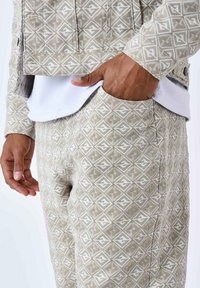 Chaqueta y pantalones beige con un patrón de diseños geométricos. La tela tiene un acabado texturizado. Camiseta blanca visible debajo de la chaqueta.