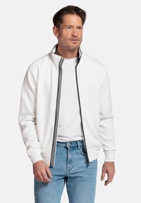 Witte ritsjack met zwarte gestreepte accenten, ribgebreide manchetten en een hoge kraag, gecombineerd met lichtblauwe jeans. Zachte textuur.