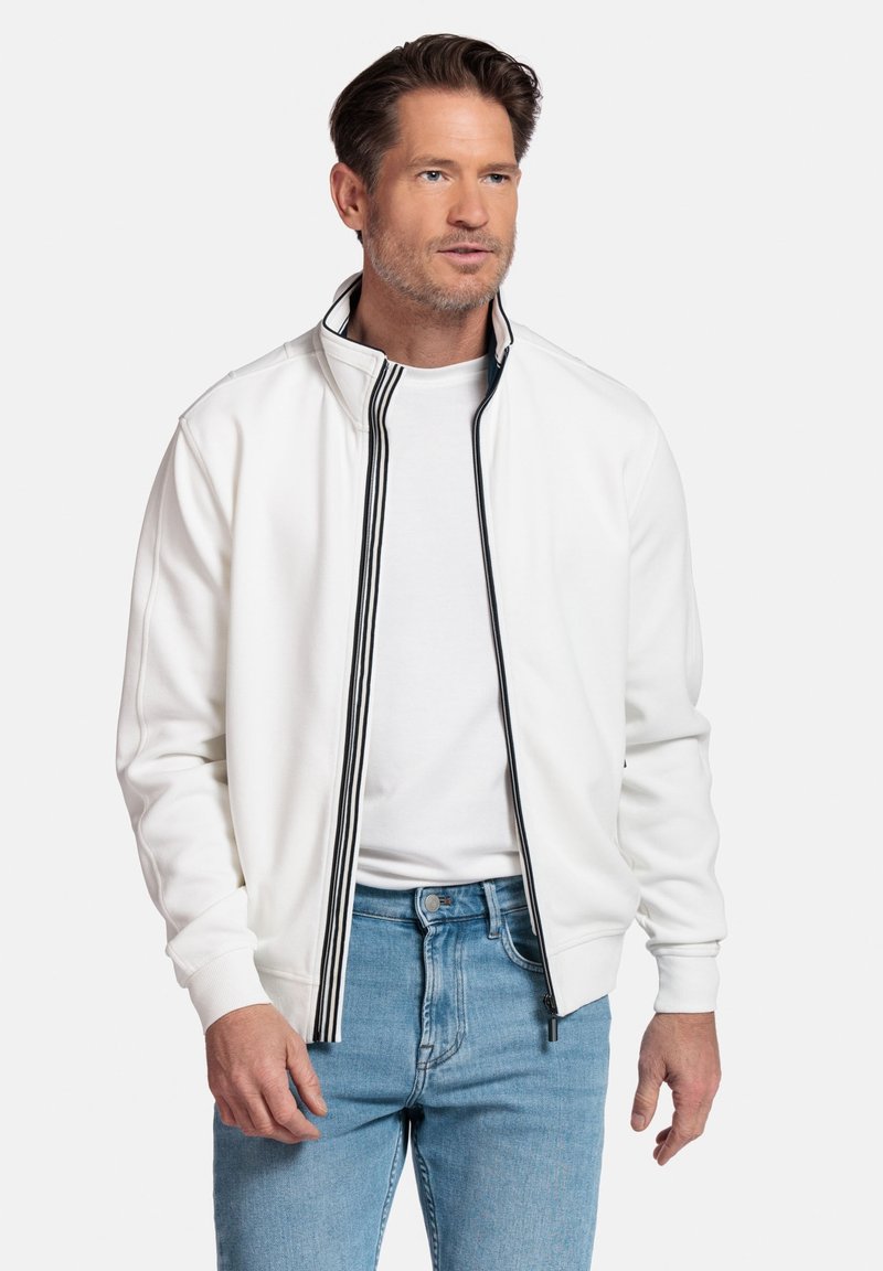 Witte ritsjack met zwarte gestreepte accenten, ribgebreide manchetten en een hoge kraag, gecombineerd met lichtblauwe jeans. Zachte textuur.