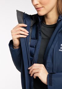 Femme en train de zipper la couche intérieure d'une veste isolante bleu marine sur un t-shirt noir, mettant l'accent sur les mains et les détails de la veste.