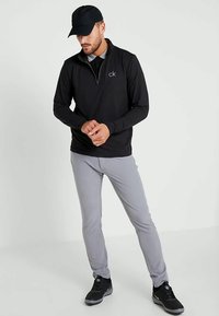 Polo preto de mangas longas com fecho de meia-zíper e logótipo, combinado com calças slim-fit cinza claro e ténis pretos, contra um fundo neutro.