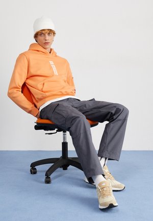 Orange hoodie med frontlomme og logo, grå cargobukser, hvid hue og beige sneakers, siddende på en orange og sort kontorstol.