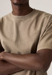 Person med mørk hudtone iført en beige Levi's t-skjorte med armene krysset foran en nøytral bakgrunn.