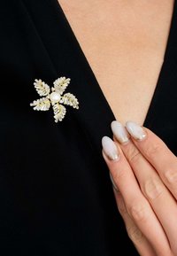 Gold and crystal floral brooch with a central pearl, featuring detailed leaf patterns, worn on a black garment. Elegant and shimmering design.