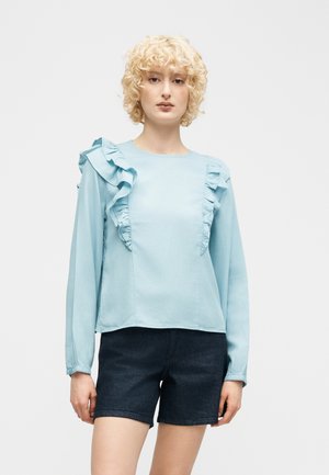 Vrouw met krullend blond haar, gekleed in een lichtblauw blouse met lange mouwen en ruches, gecombineerd met donkere denim shorts, staand tegen een effen achtergrond.