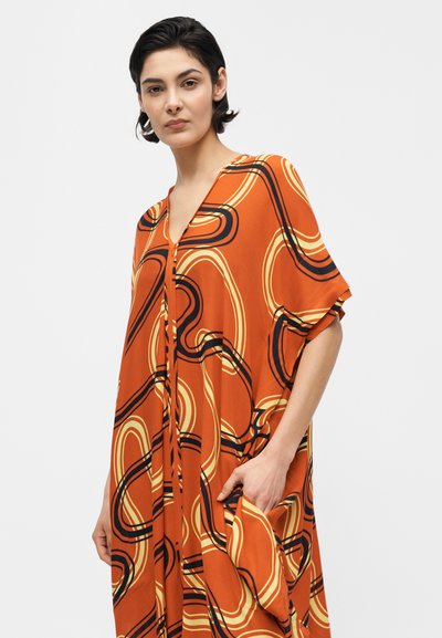 Γυναίκα με κοντά σκούρα μαλλιά, ντυμένη με πορτοκαλί φόρεμα με αφηρημένα κίτρινα και μαύρα καμπυλωτά σχέδια, με το ένα χέρι στην τσέπη.