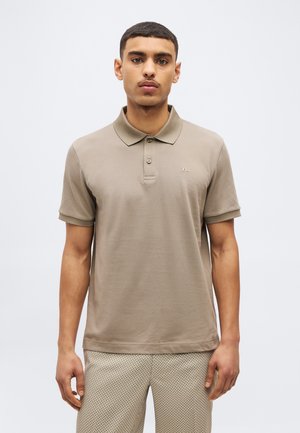 Muž stojaci čelom vpredu, oblečený v taupe krátky rukáv polo košeľu a vzorované béžové nohavice na jednoduchom pozadí.