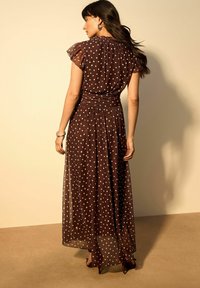 Abito lungo marrone con pois, maniche a sbuffo, vita stretta e gonna fluente. Tessuto trasparente che lascia passare la luce.